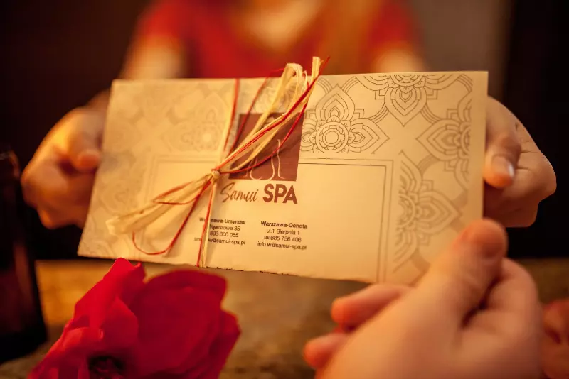 Kobieta przekazuje voucher spa do rąk klienta. Elegancji voucher z logo Samui Spa przewiązany jest wstążką. Na ladzie recepcji leżą różowe kwiaty.