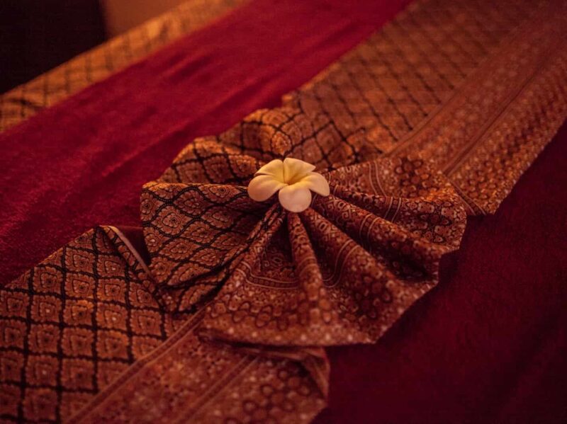 Pokój zabiegowy przygotowany do wykonania masażu aromaterapeutycznego. Na łóżku umieszczone są bordowe ręczniki oraz złote sarongi. W środku położony kwiat Frangipani.