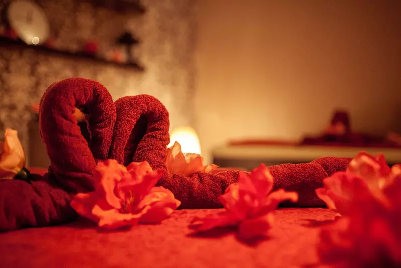 Łóżko przygotowane do wykonania romantycznego masażu dla dwojga. Na łóżku umieszczone są bordowe ręczniki ułożone w kształt serca. Wokół nich różowe kwiaty.