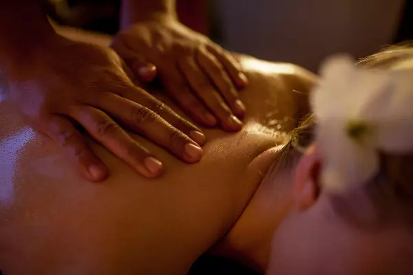 Dłonie terapeutki są ułożone na plecach klientki. Masażystka przygotowuje się do wykonania peelingu całego ciała i masażu. Kobieta ma we włosach kwiat.